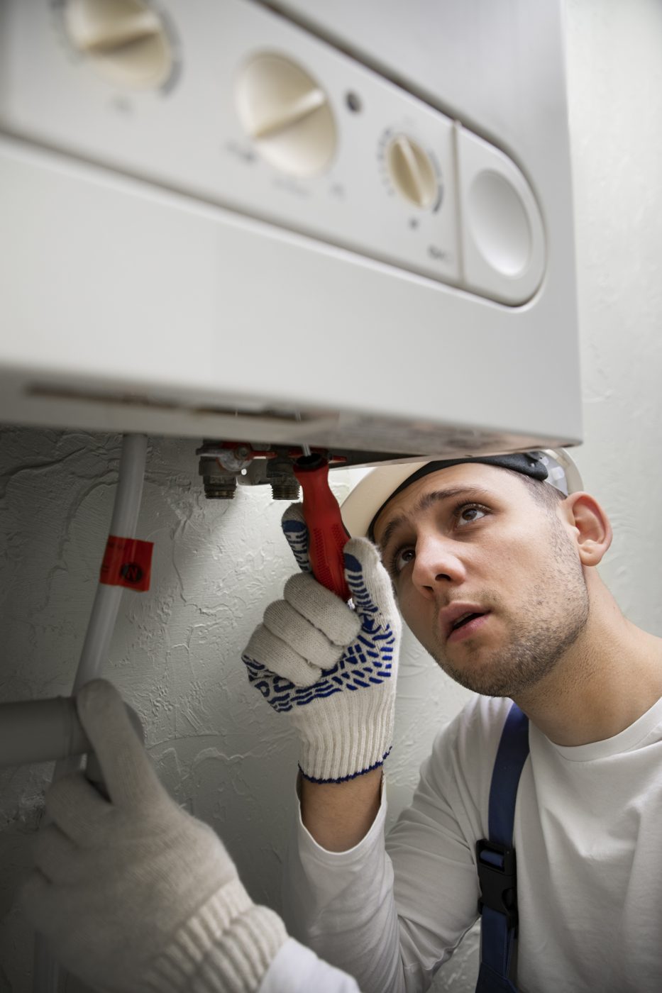 Engineer in work gloves checking the pipework beneath a wall-mounted boiler